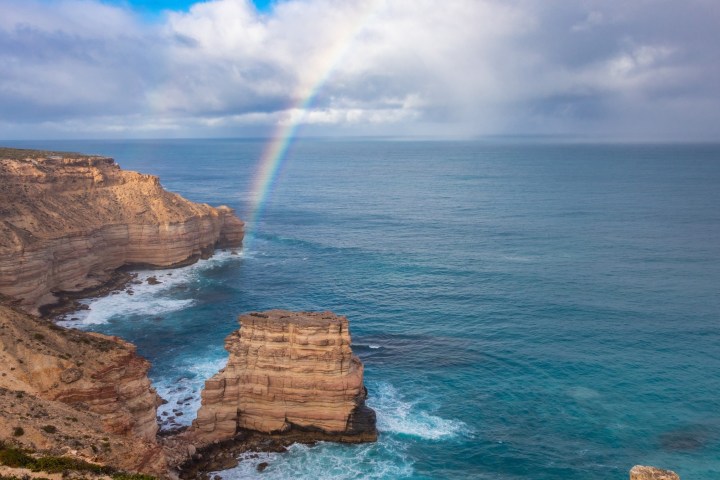 Coastal cliffs with a rainbow over the ocean under a cloudy sky.