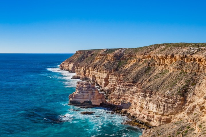 Cliffside view of ocean waves crashing against rocky cliffs under a clear blue sky.