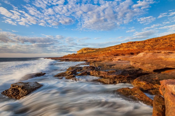 Rocky coastline at sunset with waves crashing and a cloudy sky.
